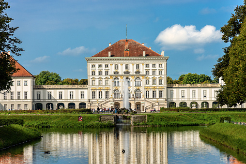 Munich, Germany - September 8th, 2018: The Nymphenburg Palace (Schloss Nymphenburg - Castle of the Nymphs) with the reflection in the river. The palace was the main summer residence of the former rulers of Bavaria of the House of Wittelsbach. The palace was commissioned by the prince electoral couple Ferdinand Maria and Henriette Adelaide of Savoy to the designs of the Italian architect Agostino Barelli. A group of tourists are visiting the ancient palace