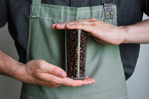 A glass beaker with coffee beans in the hands of a barista. Ready to brew roasted coffee. A man in a culinary apron. Fresh organic coffee. Disposable paper cup. Making coffee, professional service