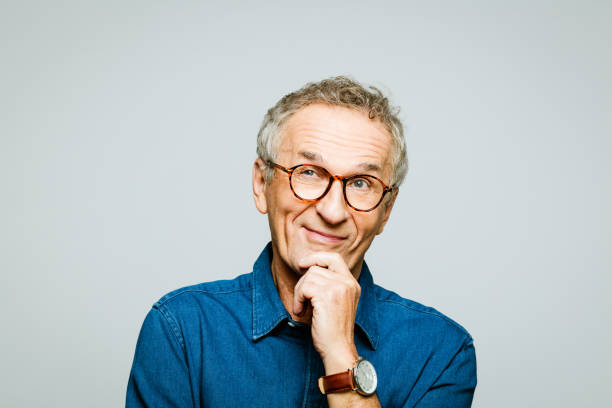 Headshot of senior man smirking with hand on chin Portrait of elderly man wearing white denim shirt and glasses looking away and smiling with hand on chin. Pleased senior entrepreneur, studio shot against grey background. curiosity stock pictures, royalty-free photos & images