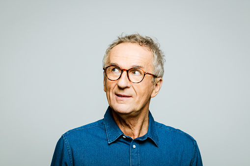 Portrait of elderly man wearing white denim shirt and glasses looking away. Thoughtful senior entrepreneur, studio shot against grey background.