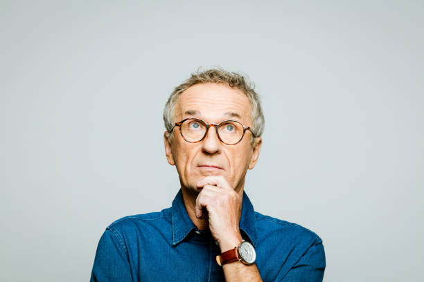 Headshot of thoughtful senior man Portrait of elderly man wearing white denim shirt and glasses looking up with hand on chin. Thoughtful senior entrepreneur, studio shot against grey background. asking yourself stock pictures, royalty-free photos & images