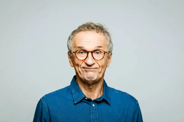 Portrait of elderly man wearing white denim shirt and glasses looking at camera. Displeased senior entrepreneur, studio shot against grey background.