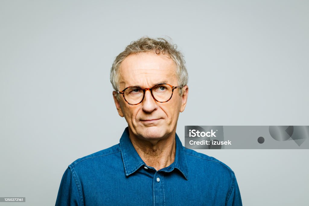 Headshot of worried senior man Portrait of elderly man wearing white denim shirt and glasses looking away. Thoughtful senior entrepreneur, studio shot against grey background. Asking Stock Photo