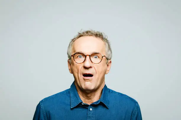 Portrait of elderly man wearing white denim shirt and glasses staring at camera with mouth open. Terrified senior entrepreneur, studio shot against grey background.