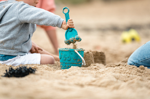 A young boy playing with a sand bucket and spade on the beach, his face is out of shot.