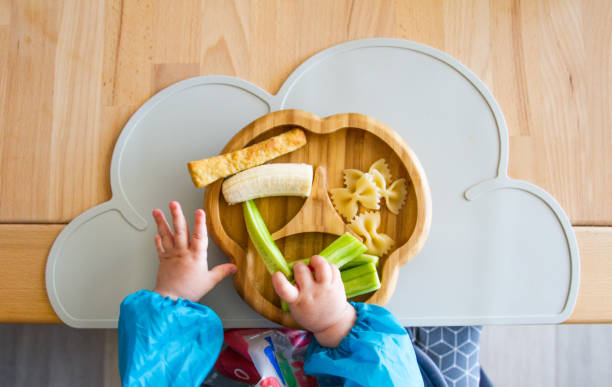 Baby nutrition BLW (baby-led weaning) with baby hands grabbing for food Baby hands grabbing food from plate (bamboo) with finger food (cucumber, pasta, banana) for babies in top view, showing the concept of baby-led weaning, nutrition without pureed food healthy eating in babies stock pictures, royalty-free photos & images