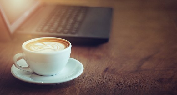 close up latte art coffee in cup on wooden table with vintage light and blur laptop on background