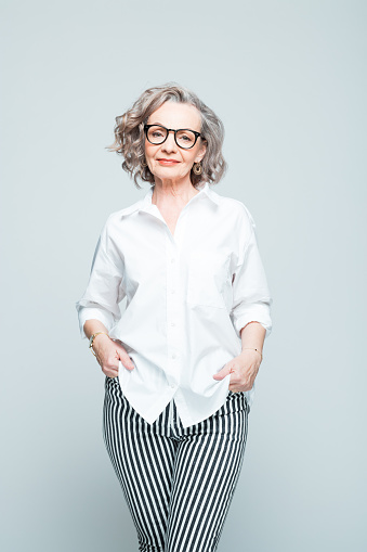 Elderly lady wearing white shirt, striped trousers and glasses standing against grey background, smiling at camera. Studio shot of female designer.