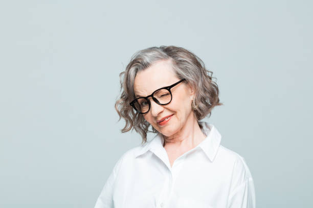 Headshot of cheerful senior woman in white shirt Elderly lady wearing white shirt and glasses standing against grey background, blinking an eye. Studio shot of female designer. blinking stock pictures, royalty-free photos & images