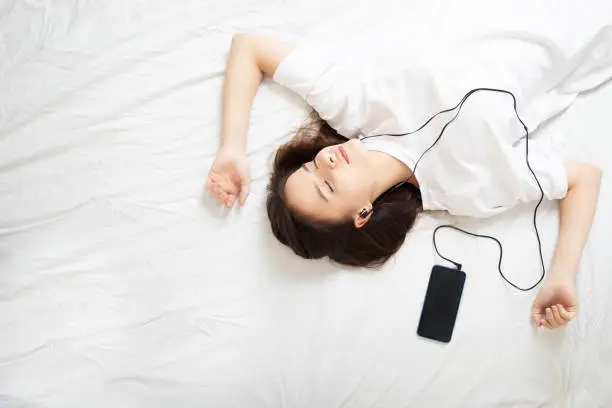 Photo of The girl lies on the bed with a smartphone, listens to an audio book with his eyes closed