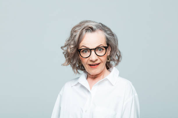Headshot of surprised senior woman in white shirt Elderly lady wearing white shirt and glasses standing against grey background, staring at camera with mouth open. Studio shot of female designer. rolling eyes stock pictures, royalty-free photos & images