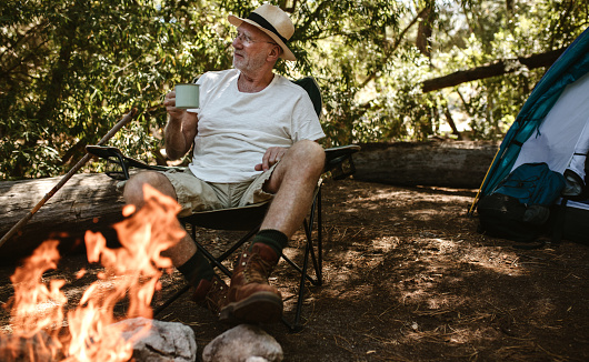 Retired man sitting near campfire and having coffee. Senior male camping in forest having a refreshing coffee in morning.