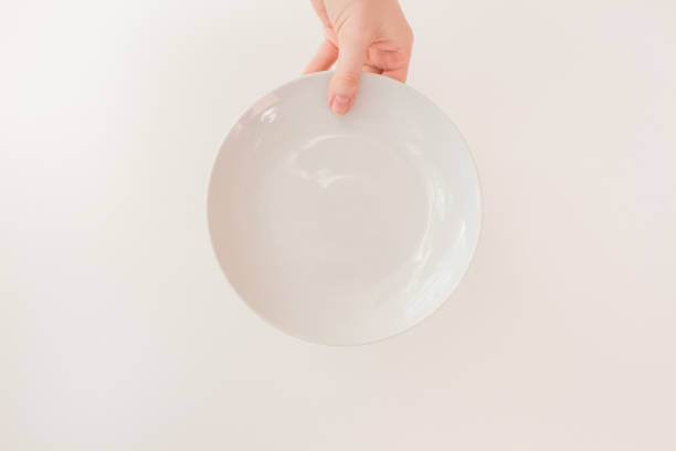 istock Female hand holds a white round plate on a white background as a mockup with copy space. Ceramic flat saucer 1250290049