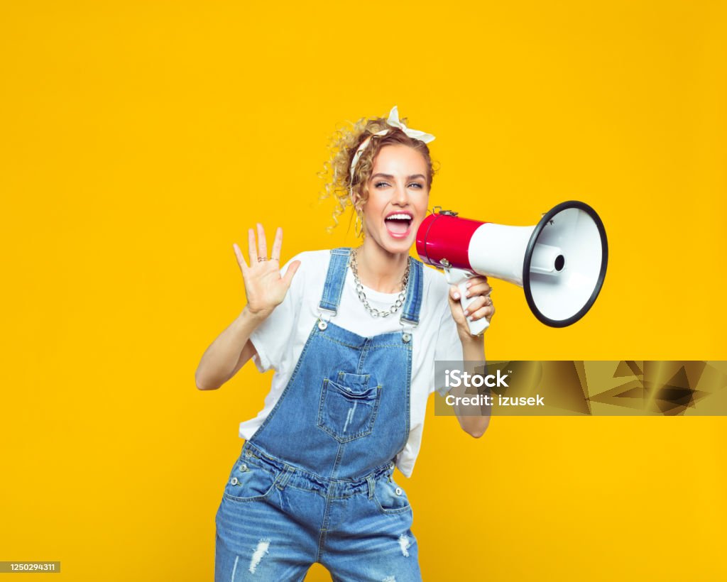 Young woman in dungarees shouting into megaphone Portrait of excited young woman wearing white t-shirt, denim dungarees and bandana shouting into megaphone. Studio shot on yellow background. Megaphone Stock Photo
