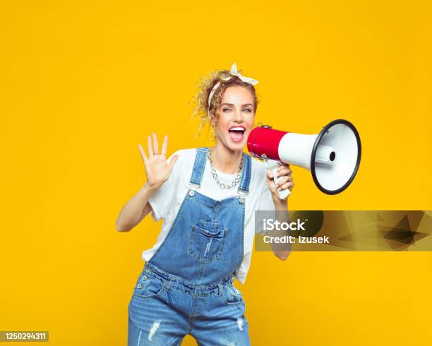 Young Woman In Dungarees Shouting Into Megaphone Stock Photo - Download Image Now - Megaphone, Colored Background, Women