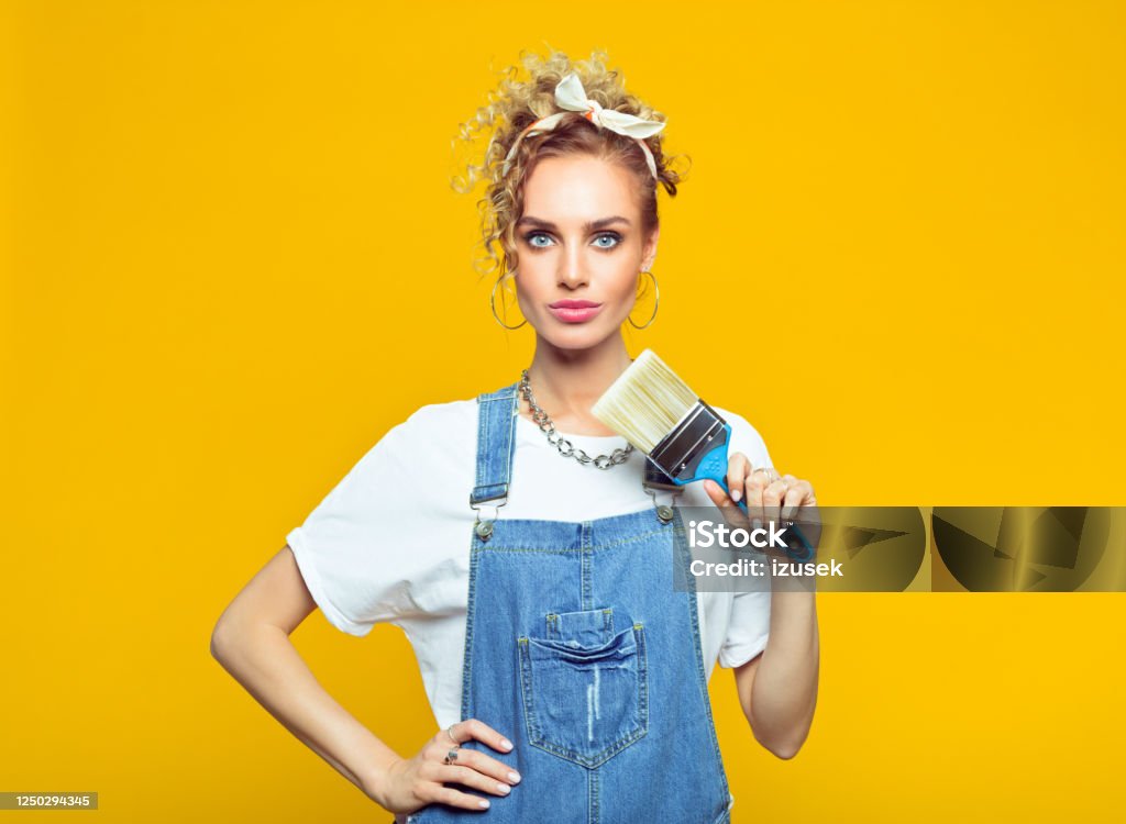 Young woman in coveralls holding paintbrush Portrait of confident young woman wearing white t-shirt, denim dungarees and bandana holding paintbrush, looking at camera. Studio shot on yellow background. House Painter Stock Photo