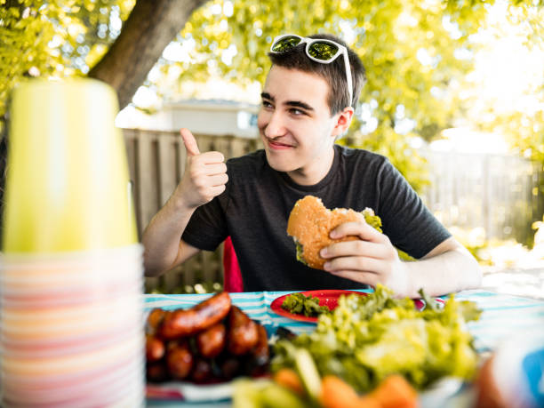 Teenage boy enjoying BBQ grill lunch outdoor at 4th July backyard celebration. Thumbs up for a hamburger.