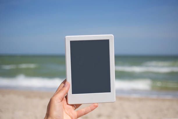 istock Reading during vacation, nice leisure, female hand hold square e-book reader tablet in white frame with dark grey screen 1250305281