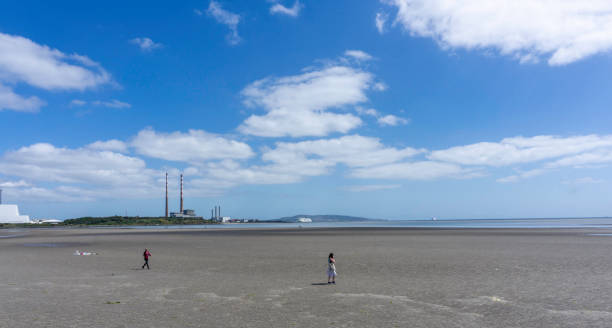 people walking along Sandymount Strand in Dublin Ireland. people walking along Sandymount Strand in Dublin with the twin towers of Poolbeg in the distance and a car ferry entering Dublin Port. bloomsday stock pictures, royalty-free photos & images