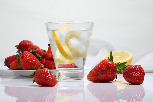 Glass of water with ice and lemon, strawberry berries on a white glossy table