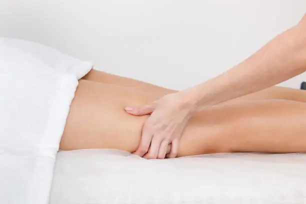 Photo of Kneading with a massage of the muscles of the legs on the hips of a model lying on a couch against a white wall.