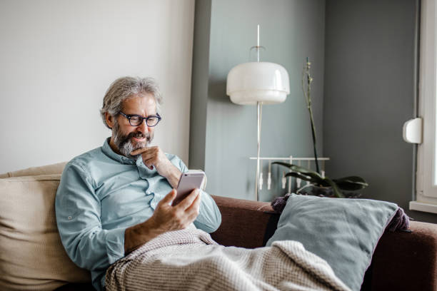 Mature man is using mobile phone at home A mature man is in her modern apartment using a mobile phone demanding photos stock pictures, royalty-free photos & images