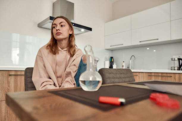 Another Perspective. Young caucasian woman looking aside while smoking marijuana from a bong or glass water pipe, sitting in the kitchen. Red marijuana grinder and lighter on the table Young caucasian woman looking aside while smoking marijuana from a bong or glass water pipe, sitting in the kitchen. Red marijuana grinder and lighter on the table. Dependence on light drugs concept pitter stock pictures, royalty-free photos & images