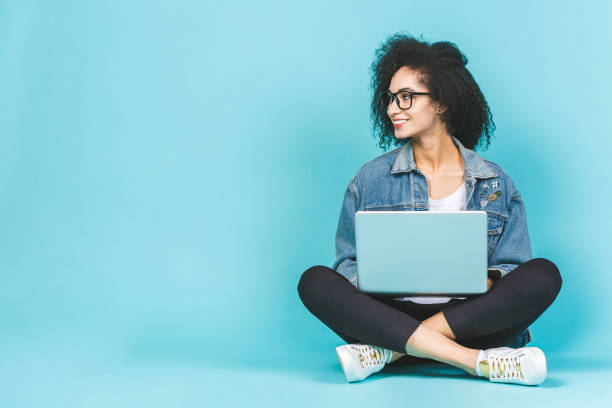 porträt einer lächelnden jungen afro amerikanischen frau mit laptop, während auf einem boden mit beinen über blauen hintergrund gekreuzt sitzen. - floor women sitting yoga stock-fotos und bilder