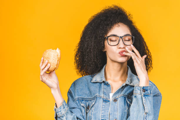 노란 배경에 고립 된 햄버거를 먹는 아프리카 계 미국인 흑인 젊은 여성. - hamburger bun bread isolated 뉴스 사진 이미지