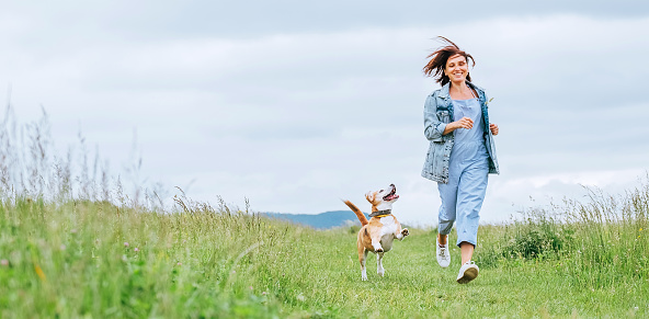 Happy smiling jogging female with fluttering hairs and her beagle dog running and looking at eyes. Walking by meadow grass path in nature with pets, healthy active people lifestyle concept image.