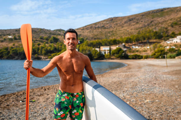 Front view of Spanish paddleboarder in early 40s standing on beach and smiling at camera before exercising in Mediterranean.