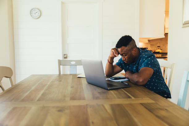 Worried Man Doing Finances at Home A 30 year old man works on his finances at his kitchen table. He is working on his bank account numbers and preparing to pay his taxes. Times are tough and making ends meet is difficult. unemployment stock pictures, royalty-free photos & images