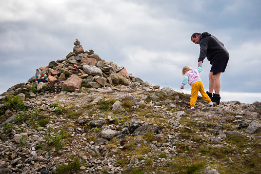 Aberdeenshire, Scotland, UK - June 22, 2019: A father holds the hand of his young daughter as they climb up a steep hill in the rocky landscape of Cairn o Mount, 1493 feet tall, on a cold windy day, Grampian Mountains, Aberdeenshire, Scotland, UK, Europe