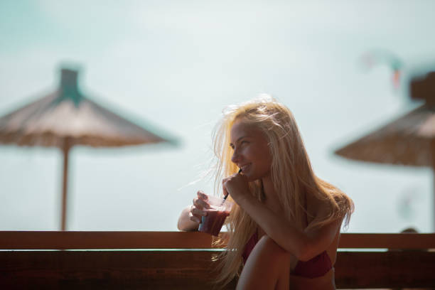 istock Young attractive blonde in bikini sitting on bench on beach and drinking refreshment. 1250402366