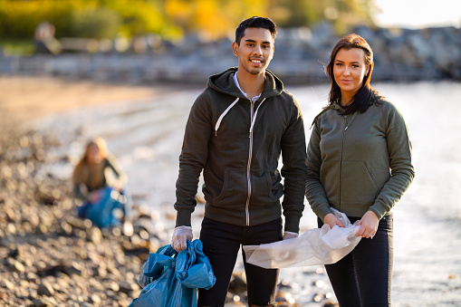 Portrait of serious multi-ethnic volunteers standing at beach. Confident young man and woman are cleaning on sunny day. They are wearing casuals.