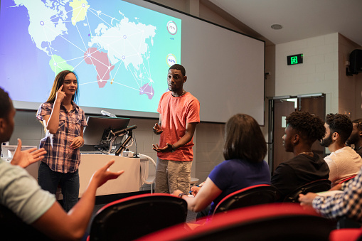 A shot of a group of students at a lecture at a university in Perth, Australia. One of the students is living with hearing impairment is presenting a slideshow using sign language.