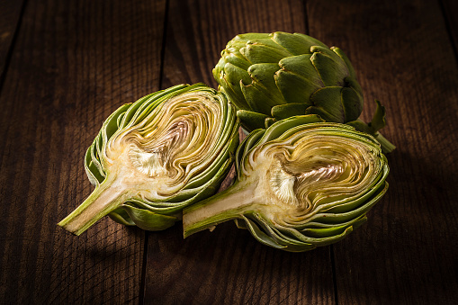 Front view of a whole and a sliced artichoke isolated on a wooden plank. Predominant colors are brown and green. Low key DSLR photo taken with Canon EOS 6D Mark II and Canon EF 24-105 mm f/4L