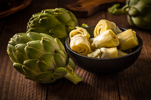 Front view of a black bowl full of artichoke hearts surrounded by two whole artichokes on a wooden plank. Predominant colors are brown and green. Low key DSLR photo taken with Canon EOS 6D Mark II and Canon EF 24-105 mm f/4L