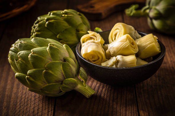 Artichoke hearts on a black bowl surrounded by artichokes Front view of a black bowl full of artichoke hearts surrounded by two whole artichokes on a wooden plank. Predominant colors are brown and green. Low key DSLR photo taken with Canon EOS 6D Mark II and Canon EF 24-105 mm f/4L artichoke stock pictures, royalty-free photos & images