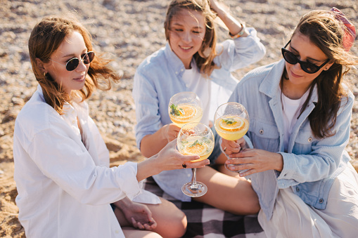 Three young beautiful happy women female friends having cozy summer picnic with lemonade, fresh bread and fruits on a beach at sunset.