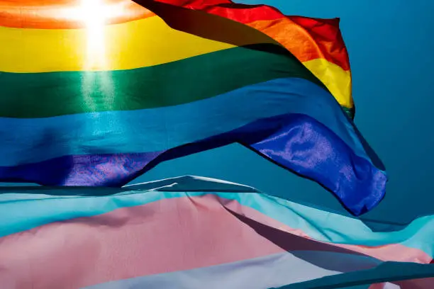 closeup of a gay pride flag and a transgender pride flag waving on the blue sky, moved by the wind, with the sun in the background