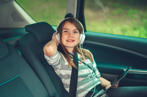Shot of a young girl sitting in a car wearing headphones and using a digital tablet