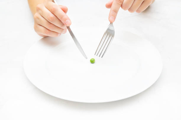 Female hands holding a knife and a fork over a small pea on a large white plate. Extreme dieting and eating disorder concept. big plate of food stock pictures, royalty-free photos & images