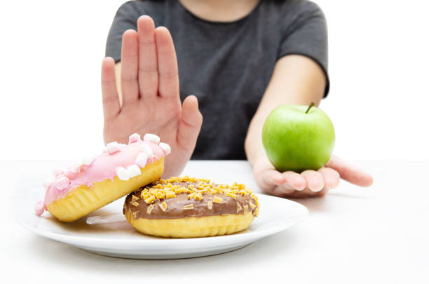 Woman choosing a fresh apple over a plate of donuts, rejecting junk unhealthy food with a hand gesture. Closeup, selective focus. Dieting and healthy eating concept. avoidance stock pictures, royalty-free photos & images