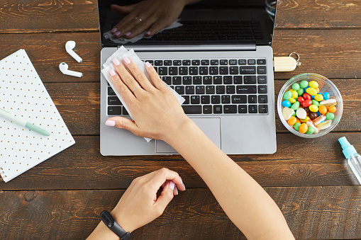 Top down view at unrecognizable young woman sanitizing laptop with cleaning wipe while working at home office, copy space