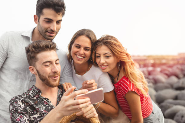 istock Young friends sitting outdoors and looking at smartphone. Young man showing something interesting to friends outdoors at the seaside 1250440563