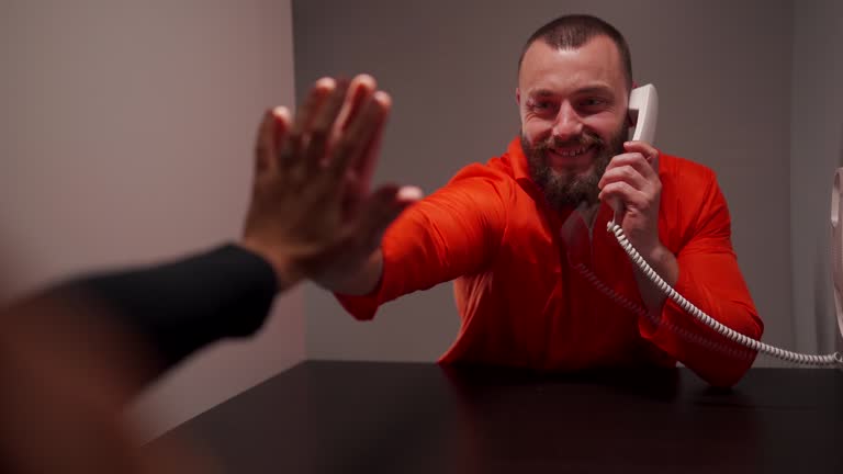 Prisoner in jumpsuit and his wife putting hands on glass between them during visit at prison