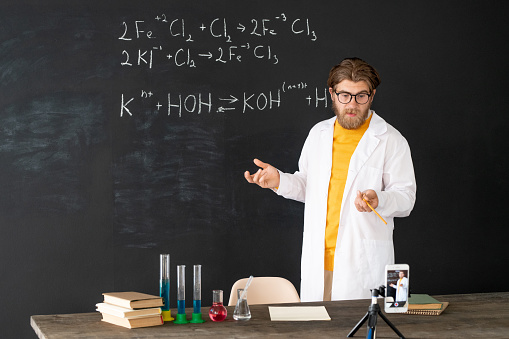 Contemporary teacher in whitecoat doing record of chemistry lesson for his online audience on smartphone camera against blackboard