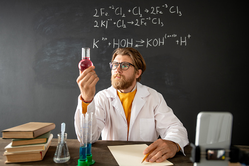 Bearded teacher of chemistry looking at tube with pink liquid substance while sitting by table in front of smartphone camera at lesson