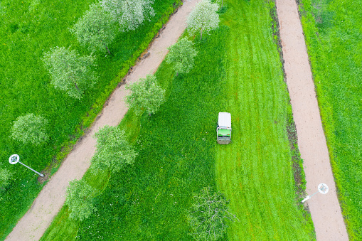 Lawn mower machine rides on grass, mowing tall grass in a city park among trees and walking paths, aerial top view